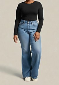 Femme portant un haut noir à manches longues, un jean bleu clair taille haute à jambes larges, et des chaussures blanches, debout devant un fond beige uni.