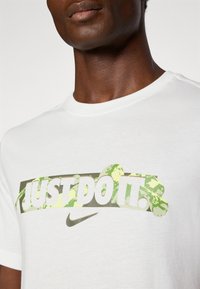 Biele bavlnené tričko s nápisom "JUST DO IT." v zelenom maskáčovom vzore, s logom Nike. Klasický okrúhly výstrih a štandardné krátke rukávy.