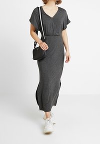 Grau gesprenntes Maxikleid mit V-Ausschnitt, kurzen, schwingenden Ärmeln, seitlichen Schlitzen und elastischem Bund. Kombiniert mit einer kleinen schwarzen Umhängetasche und Sneakern.