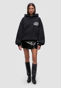 Czarna, przylegająca bluza z kapturem z przednią kieszenią oraz białym napisem na klatce piersiowej, połączona z błyszcząca, czarną mini spódnicą i czarnymi botkami do kolan.