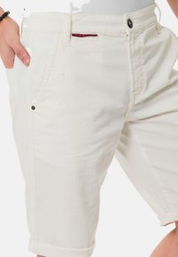 Shorts pour homme de couleur crème en coton, dotés d'un système de fermeture à bouton, de poches latérales et d'ourlets roulés; texture douce et détails minimalistes.