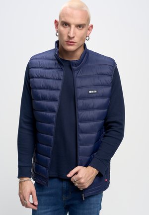 Navy Puffer-Weste mit horizontalem Steppmuster, Frontreißverschluss undLogopatch. Getragen über einem navyblauen Strickpullover und blauen Jeans.