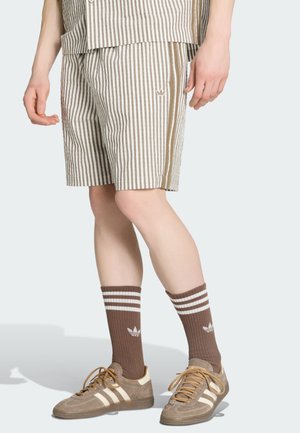 Piernas con pantalones cortos a rayas marrón y blanco, calcetines marrones a juego con rayas blancas y zapatillas marrón claro con detalles blancos.