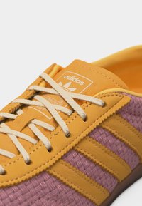 Κίτρινο και μοβ αθλητικό παπούτσι Adidas με λευκά κορδόνια, με το λογότυπο της Adidas στη γλώσσα και υφασμάτινες πλαϊνές επιφάνειες με υφή.