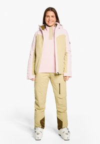 Ski outfit with a pink and beige insulated jacket, beige pants with side pockets, and white ski boots featuring gray accents.