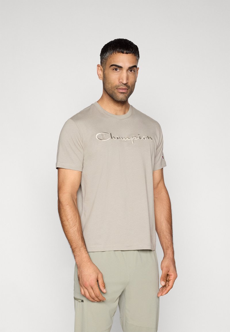 Μπεζ βαμβακερό t-shirt με απαλή υφή, κοντά μανίκια και κεντημένο λογότυπο "Champion" στο μπροστινό μέρος. Συνδυάζεται με ανοιχτόχρωμο παντελόνι.