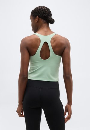 Vrouw met gevlochten haar, gekleed in een lichtgroen racerback tanktop en zwarte legging, staand met de rug naar de kijker tegen een effen achtergrond.