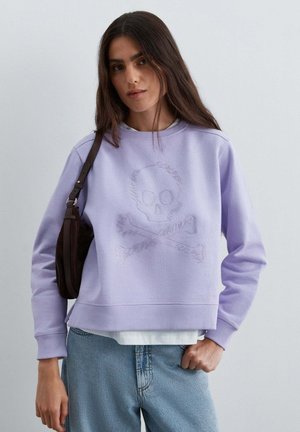 Mujer con sudadera lavanda con un diseño sutil de calavera y huesos cruzados, vaqueros azul claro y un bolso oscuro de hombro.