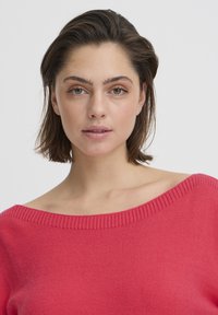 Pull rose avec un large col côtelé. Texture douce et coupe décontractée. Cheveux portés lâchés. Fond neutre.