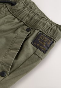 Shorts cargo vert olive avec une ceinture élastique, des poches avant, un bouton pression noir et une étiquette indiquant les coordonnées et le numéro d'édition.