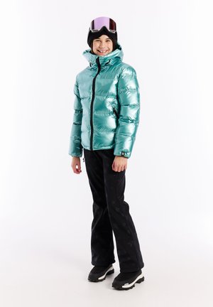 Metallisch türkiser Pufferjacke mit hohem Kragen, schwarzem Reißverschluss und Seitentaschen, kombiniert mit schwarzen, sternmusternden Hosen und Winterstiefeln.