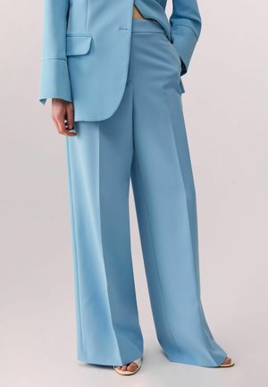 Persona che indossa pantaloni larghi azzurri chiari e blazer abbinato, con una mano in tasca e sandali bianchi aperti sulle dita visibili.