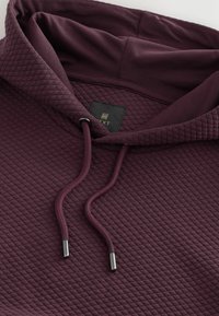 Burgunderfarbene Kapuzenjacke mit gestepptem Design, verstellbaren Zugbändern mit metallischen Spitzen und einem Etikett mit der Aufschrift "NEXT" im Kragen.
