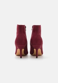 Botas de tobillo de ante burdeos con un diseño elegante, que presentan una punta afilada, un alto tacón de aguja y cierre de cremallera en la parte posterior.