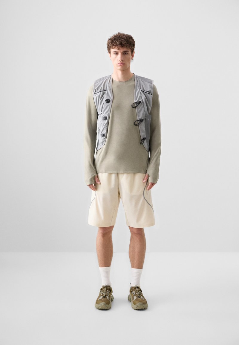 Grå vest med knapper over en lys grøn langærmet skjorte, parret med kridhvide shorts og olivengrønne sneakers. Enkel design, funktionelle egenskaber.