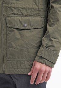 Olive green lightweight jacket featuring a front flap pocket, one snap closure, and a textured surface with quilted stitch detailing.