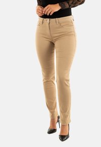 Donna che indossa pantaloni beige aderenti, top con maniche in pizzo nero e scarpe con tacco nere, in piedi su uno sfondo bianco.