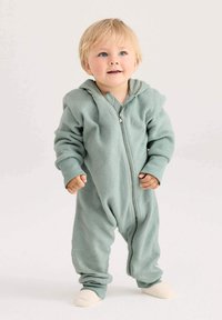 Peuter met blond haar, gekleed in een lichtgroene fleece zip-up romper, staande voor een neutrale achtergrond en iets omhoog kijkend.