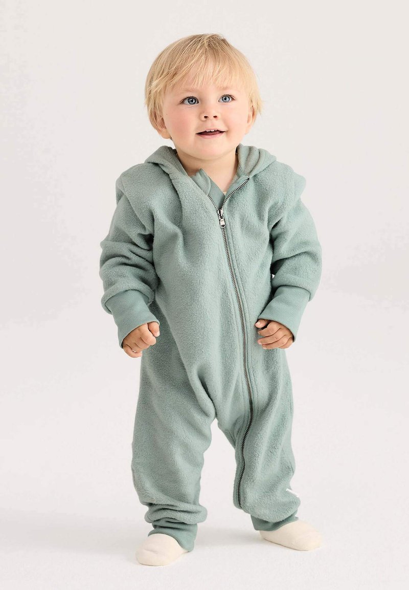 Peuter met blond haar, gekleed in een lichtgroene fleece zip-up romper, staande voor een neutrale achtergrond en iets omhoog kijkend.