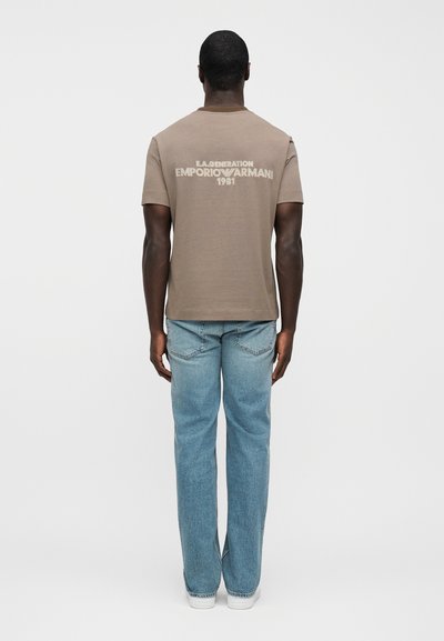 Καφέ κοντομάνικο μπλουζάκι με λευκό κείμενο "E.A. Generation Emporio Armani 1981" στην πλάτη, συνδυασμένο με ανοιχτό μπλε τζιν και λευκά παπούτσια.