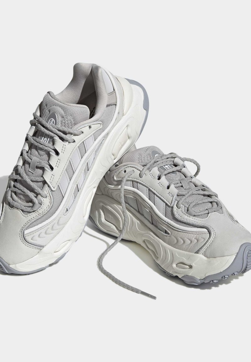 Hellgraue Sportschuhe mit einem Obermaterial aus Mesh und Synthetik, einem abgerundeten Design, einer dicken Sohle und strukturierten Akzenten mit Schnürsenkeln.