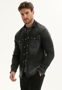 Donkergrijze spijkerblouse met een klassiek ontwerp, voorzien van twee borstzakken, drukknoopsluitingen en contrasterende stiksels. Zachte textuur en aansluitende pasvorm.