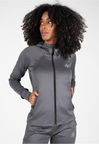 Grijze sportieve zip-up jas met een capuchon, gemaakt van gladde stof. Voorzien van ritszakken en een logo-accent op de borst.