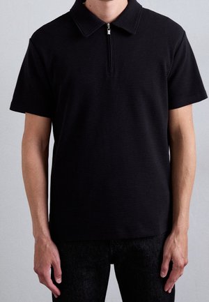 Personne portant un polo noir à manches courtes avec un col zippé à l'avant, debout, les bras relâchés le long du corps.