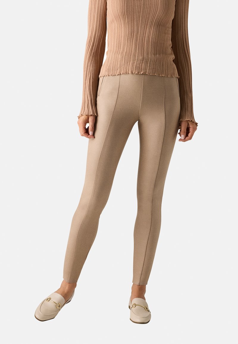 Leggings beige con una textura suave, que presentan costuras verticales y un ajuste ceñido. Combinados con una blusa de manga larga acanalada y mocasines.