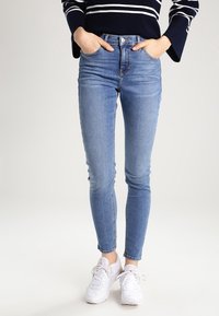 Jeans ajustados de mezclilla azul desgastada, de cintura alta con un diseño clásico de cinco bolsillos, combinados con zapatillas deportivas blancas y una blusa de rayas en color marino.