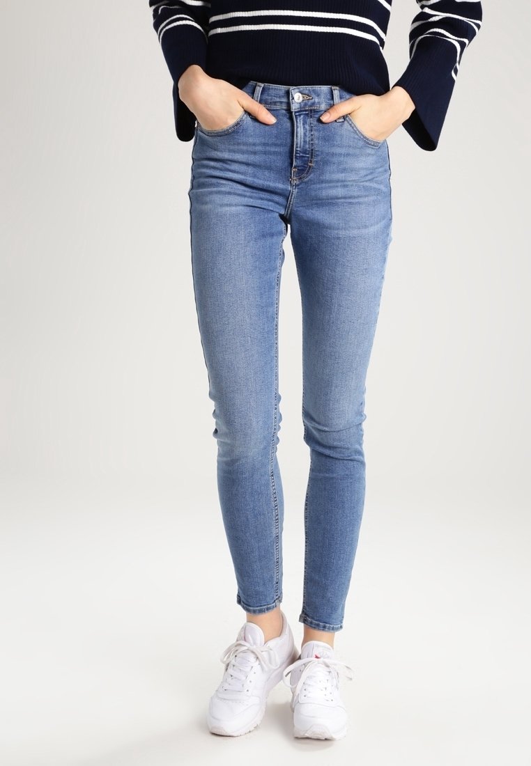 Jeans ajustados de mezclilla azul desgastada, de cintura alta con un diseño clásico de cinco bolsillos, combinados con zapatillas deportivas blancas y una blusa de rayas en color marino.