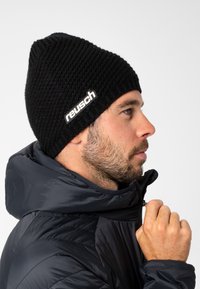 Black knitted beanie with a textured pattern and a white "reusch" logo on the side, worn by a person in a dark puffer jacket.