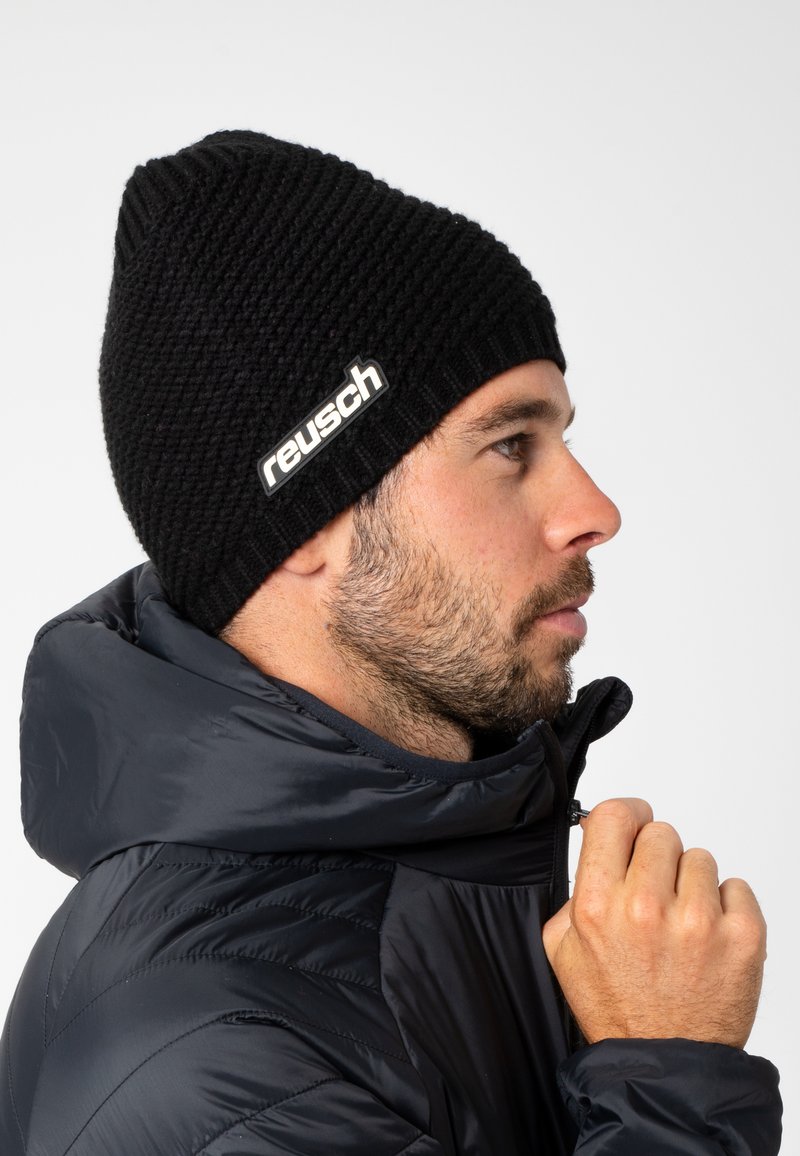 Black knitted beanie with a textured pattern and a white "reusch" logo on the side, worn by a person in a dark puffer jacket.