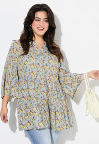 Blomster tunika i pastelfarver med lagdelt design, V-udskæring og flagrende ærmer. Stylet med lyse denimbukser og en hvid håndtaske.