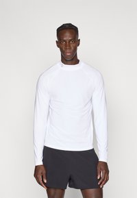 Fehér, hosszú ujjú sportpóló raglán ujjakkal, sima felülettel és egy finom logóval a galléron, fekete rövidnadrággal párosítva.