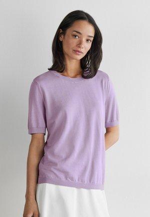 Femme aux cheveux bruns mi-longs, portant un haut à manches courtes violet clair et une jupe blanche, debout devant un mur blanc uni.