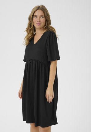 Femeie cu păr blond ondulat, purtând o rochie neagră largă cu decolteu în V și mâneci scurte, stând într-un fundal simplu și luminos.