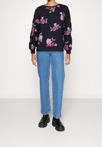 Personne portant un pull marine avec des imprimés floraux roses, un jean bleu clair à jambes droites et des bottines plateformes noires épaisses.