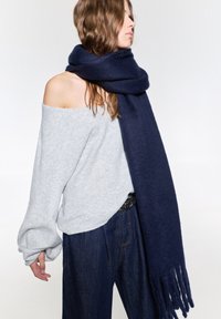 Marineblå uldhalstørklæde med frynser, draperet løst omkring halsen. Grå off-shoulder sweater og brede blå jeans fuldender looket.