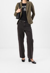Olijfblazer over een zwart graphic t-shirt, gecombineerd met hoge zwarte jeans en zwarte lakleren hakken. Zwarte handtas over de schouder.