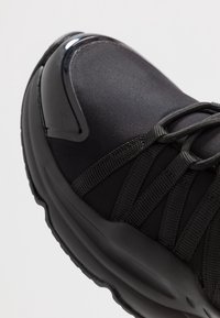 Chaussure de sport noire avec une tige synthétique lisse, des accents texturés et une semelle intermédiaire épaisse. Les lacets sont intégrés avec des sangles pour le soutien.