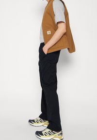 Chaleco sin mangas marrón sobre una camiseta gris, combinado con pantalones cargo negros y zapatillas de correr negras con detalles en amarillo.