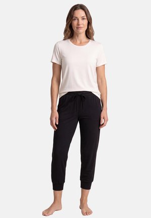 Kvinde står barfodet iført en hvid kortærmet t-shirt og sorte joggingbukser med snøre ved livet mod en ensfarvet baggrund.