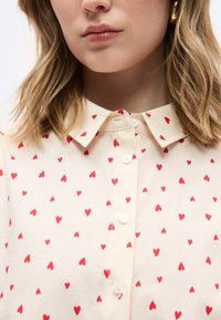 Γυναίκα με ξανθά μαλλιά που φοράει πουκάμισο κρεμ με κουμπιά, διακοσμημένο με μικρά κόκκινα μοτίβα καρδιών.