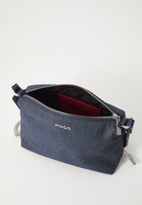 Marine denim crossbody-veske med sølvtilbehør, glidelås-lukking og mørkt interiør. Har en synlig logo og et rødt kort inni.