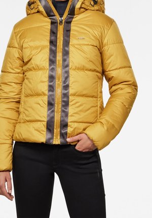 Personne portant une veste matelassée jaune moutarde avec une fermeture éclair grise brillante et un pantalon noir, main dans la poche, sur fond blanc.