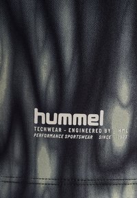 Svart och grått strukturerat tyg med ett subtilt mönster, med vit text "hummel" och detaljer om teknisk och sportkläders prestanda.