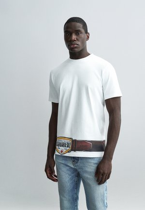 Homme portant un t-shirt blanc avec ceinture marron imprimée et motif écusson, associé à un jean bleu clair, debout devant un fond uni clair.