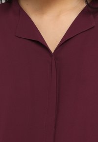 Blusa bordeaux con scollatura a V, realizzata in tessuto morbido. Presenta una cucitura sottile lungo la parte anteriore e bordi del colletto svasati.