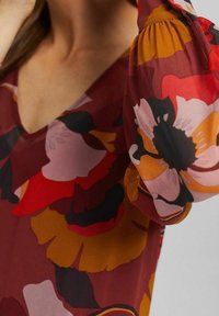 Röd blommig blus med V-ringning, utrustad med puffärmar. Tyget har stora, färgglada blommor i nyanser av röd, rosa och gul.